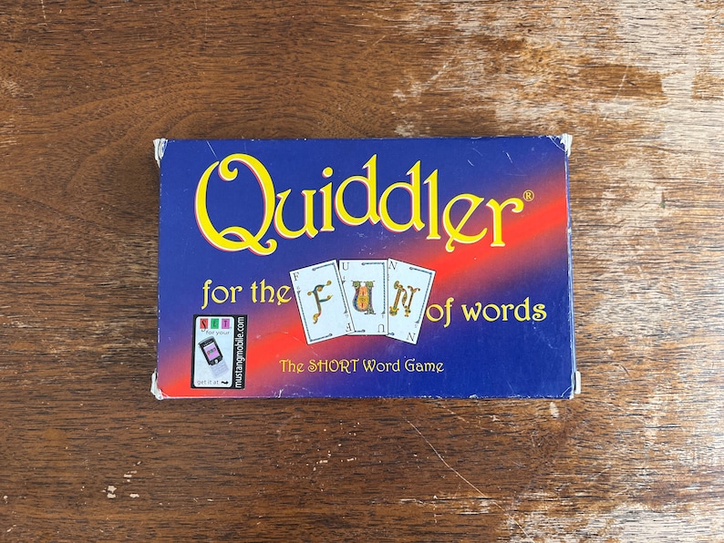 May include: A blue game box for "Quiddler" with yellow and red text. The box features the game's name, the phrase "for the FUN of words," and the tagline "The SHORT Word Game." Three playing cards are also visible.