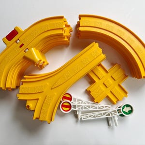 May include: A collection of yellow plastic toy train track pieces, including curved sections, a Y-shaped switch track, and a cross-shaped intersection piece, accompanied by two white plastic signal arms with red and green circular signs.