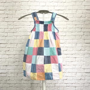 May include: A colorful patchwork dress with a square pattern in shades of pink, blue, yellow, and white. The dress has a square neckline and straps, hanging on a hanger against a white brick wall.