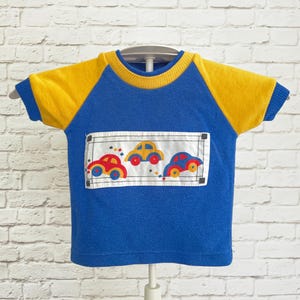 May include: A blue short-sleeve shirt with yellow raglan sleeves and a ribbed yellow collar. The center features a white rectangular panel with a smocked design depicting three small cars in red, yellow, and blue with colorful geometric accents.