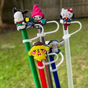 May include: Five colorful reusable straws with different cartoon character toppers. The toppers include a yellow dog, a pink Hello Kitty-like character, a white and blue character, a black and white dog, and a red, white, and blue Hello Kitty-like character.