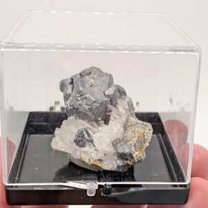 May include: A mineral specimen encased in a clear, rectangular display case. The specimen features a cluster of gray, metallic-looking minerals atop a bed of white quartz crystals. The display case sits on a black base.