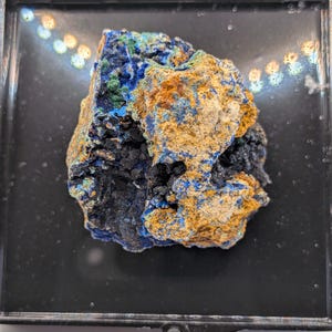 May include: A colorful mineral specimen featuring a mix of blue, green, orange, and black hues. The rough-hewn rock is displayed in a black, rectangular display case. The mineral's surface has a textured appearance.
