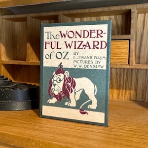 May include: A vintage book cover featuring a green background with the title "The Wonderful Wizard of Oz" in red and white lettering. The author, L. Frank Baum, and illustrator, W.W. Denslow, are listed below the title. The cover features a cartoon illustration of a lion with a crown, glasses, and a red beard.