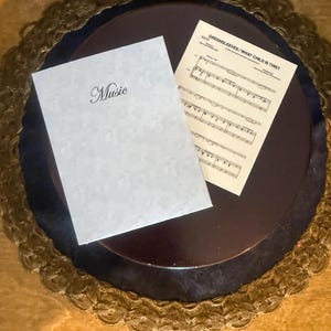 May include: A white booklet with the word "Music" in cursive script and a sheet of music titled "Greensleeves / What Child Is This?" are on a dark brown surface. The surface is surrounded by a decorative gold-colored trim.