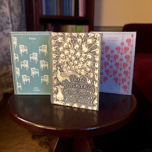 May include: Three miniature hardcover books on a dark brown wooden table. The books are "Pride and Prejudice" by Jane Austen, "Emma" by Jane Austen, and "Sense and Sensibility" by Jane Austen. The books have decorative covers with floral and peacock designs.