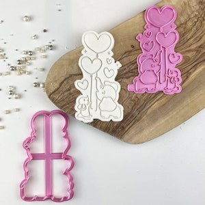 May include: Two pink plastic cookie cutters. One cutter is shaped like an elephant holding balloons in the shape of hearts. The other cutter is shaped like a stack of three hearts.