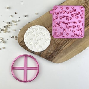 Pode incluir: Um carimbo de fondant em forma de coração rosa e um cortador redondo com um design de cruz. O carimbo está cheio de pequenos corações e o cortador tem uma moldura rosa.