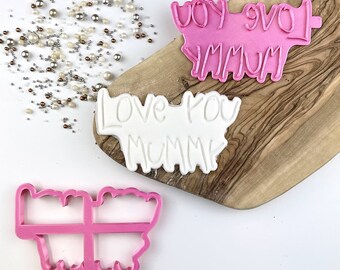 Peut inclure: Deux emportes-pièces en plastique rose, l'un en forme de papillon et l'autre avec les mots "Love You Mummy". Un emporte-pièce blanc avec les mêmes mots est également présenté sur une planche à découper en bois.