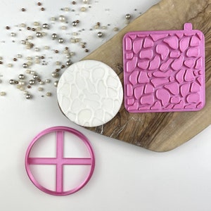 Pode incluir: Três ferramentas de decoração de bolos de plástico rosa: um cortador redondo com um design de cruz, um molde texturizado quadrado e um molde texturizado redondo.