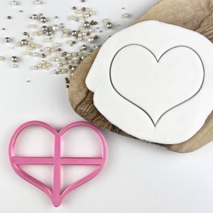 Puede incluir: Cortador de galletas de plástico rosa en forma de corazón con un diseño de rejilla cruzada dentro del corazón. El cortador está sobre una superficie blanca junto a un cortador de galletas blanco en forma de corazón sobre una tabla de cortar de madera.