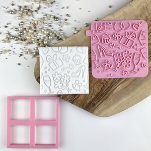 Pode incluir: Um conjunto de três formas de cozedura de silicone rosa. As formas são quadradas e apresentam uma variedade de designs com tema de casamento, incluindo um cupcake, um sapato, uma caixa de presente e uma taça de champanhe. As formas estão sobre uma superfície de madeira.