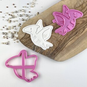 Peut inclure: Deux emportes-pièces en plastique rose et blanc en forme de dinosaures volants. Les emportes-pièces sont sur une planche à découper en bois.