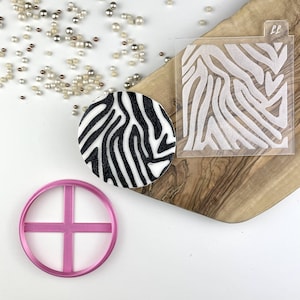 Pode incluir: Um estêncil de estampa de zebra em preto e branco, um cortador de biscoitos circular de plástico rosa e um círculo de fondant de estampa de zebra em preto e branco.