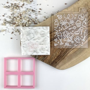 Pode incluir: Um estêncil de plástico transparente com um contorno branco de vários desenhos, incluindo uma caixa de presente, um cupcake, um sapato, uma taça de vinho e uma flor. O estêncil está sobre uma superfície de madeira com um estêncil de plástico rosa com quatro quadrados no canto inferior esquerdo.