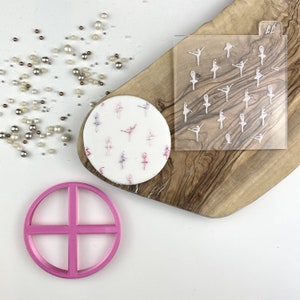 Pode incluir: Um bolo branco de fondant decorado com silhuetas de bailarinas rosa e brancas. Um estêncil de plástico transparente com silhuetas de bailarinas é mostrado ao lado do bolo. Também é mostrado um cortador de círculo de plástico rosa.