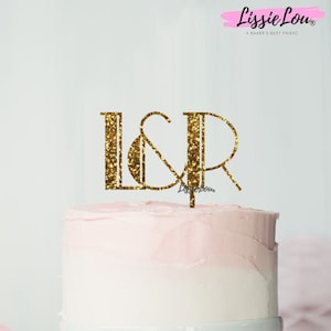 Peut inclure: Un topper de gâteau en paillettes dorées avec les lettres L & R, sur un gâteau blanc avec un glaçage rose. Le gâteau est sur un support à gâteau blanc. Le texte "Glitter Gold" est visible en bas de l'image.