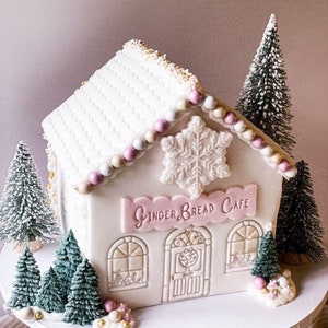 Puede incluir: Una casa de jengibre blanca decorada con detalles en rosa y dorado. La casa tiene un copo de nieve en la parte delantera y un letrero que dice "Gingerbread Cafe". Hay pequeños pinos artificiales alrededor de la casa.