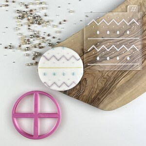 May include: A pink plastic cookie cutter shaped like a circle with a cross inside. A white cookie decorated with purple, blue, and yellow icing is next to the cutter. A clear acrylic stencil with a geometric pattern is in the background.