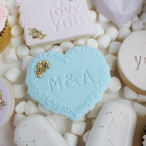 K&ouml;nnte beinhalten: Eine Sammlung dekorierter Kekse, darunter ein hellblaues herzf&ouml;rmiges Keks mit den Initialen "M&A" und einem floralen Rand. Weitere Kekse sind ein rosa Quadrat mit "love YOU" und goldenen Akzenten.