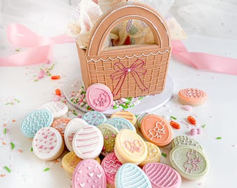 May include: A brown gingerbread basket with a pink bow and a white handle filled with pastel-colored Easter egg cookies. The cookies are decorated with different designs, including hearts, stripes, and swirls.