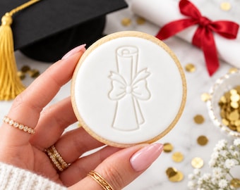 May include: A round sugar cookie with a smooth white icing surface featuring an embossed design of a rolled diploma tied with a bow. The cookie is held against a background containing a black graduation cap with a gold tassel and a scroll.