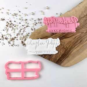 May include: Two pink and white cookie cutters with the text "Pumpkin Patch" and arrows pointing in opposite directions. The pink cutter says "This Way" and the white cutter says "Yaw Siht".