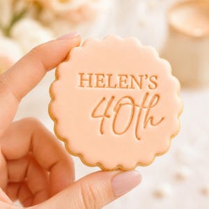 May include: A round, scalloped-edge biscuit with a smooth, pale peach-coloured icing finish. The surface features embossed text that reads "HELEN'S" in a clean, sans-serif font above the number "40th" written in a flowing, cursive script style.
