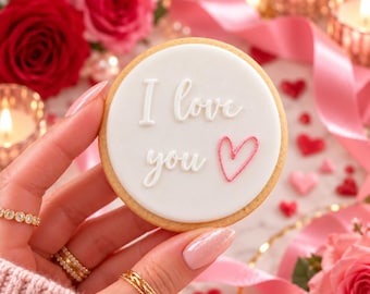 Puede incluir: Una galleta redonda con glaseado blanco liso que presenta el texto en relieve I love you en blanco y el contorno de un pequeño corazón rosa. La galleta tiene un borde dorado y se muestra ante rosas rojas y rosas con cintas.