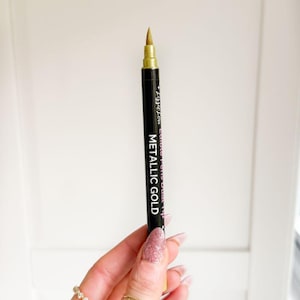 Puede incluir: Un rotulador comestible negro con una punta dorada met&aacute;lica. El cuerpo presenta texto en blanco que dice LizzeLou Edible Pens Dual Tip y METALLIC GOLD. El rotulador tiene una tapa de color dorado y una punta de fieltro para detalles dorados.
