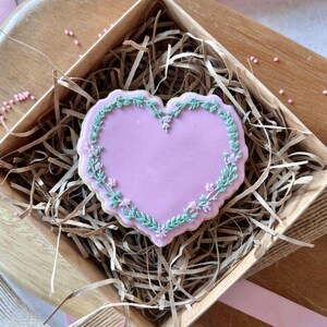 K&ouml;nnte beinhalten: Ein herzf&ouml;rmiger Keks mit hellrosa Zuckerguss in der Mitte und einem gr&uuml;n-rosa Blumenrand. Der Keks wird in einer Holzkiste mit braunen Papierschnipseln pr&auml;sentiert, was auf ein Geschenk oder einen Leckerbissen hindeutet.
