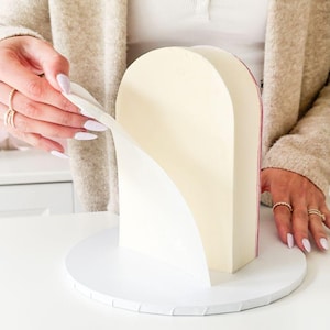 May include: A layered cake with pale yellow frosting is being revealed. The cake is in an arch shape, with white paper being peeled away. The cake sits on a white, round cake board. The background is neutral.