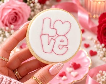 Puede incluir: Una galleta redonda con una superficie de glaseado blanco liso que presenta la palabra LOVE delineada en rosa, donde la letra O es reemplazada por una forma de corazón. La galleta tiene un borde dorado claro y una forma circular.