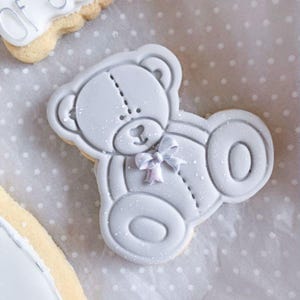 May include: A light gray teddy bear shaped sugar cookie decorated with smooth icing. The bear features a stitched line detail down the center of its head and body, a small heart shaped nose, and a delicate light blue bow tied at its neck.
