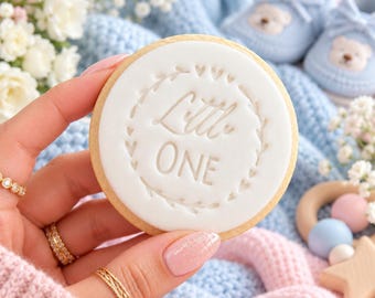 May include: A round sugar cookie topped with white fondant featuring the embossed text Little ONE surrounded by a delicate heart and leaf border. The cookie is held against a background of light blue knit fabric and baby booties.