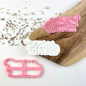 Puede incluir: Tres cortadores de galletas con la palabra BOO, una calabaza, un fantasma y un sombrero de bruja. Un cortador es rosa, otro es blanco y otro es un marco rosa. Los dise&ntilde;os incluyen l&iacute;neas en relieve detalladas para las caras y el sombrero.