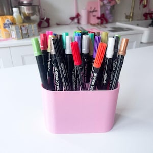 Puede incluir: Una colecci&oacute;n de marcadores comestibles de doble punta de varios colores almacenados en un recipiente de pl&aacute;stico rosa claro. Los marcadores tienen tapas de colores y cuerpos negros con texto en blanco, incluyendo etiquetas como Metallic Bronze, Hard Orange y White.
