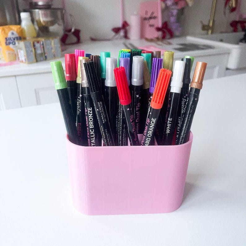 Puede incluir: Una colecci&oacute;n de marcadores comestibles de doble punta de varios colores almacenados en un recipiente de pl&aacute;stico rosa claro. Los marcadores tienen tapas de colores y cuerpos negros con texto en blanco, incluyendo etiquetas como Metallic Bronze, Hard Orange y White.