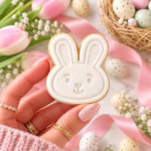 May include: A white, rabbit-shaped sugar cookie with embossed facial features, including ears, eyes, and a heart-shaped nose. The cookie has a smooth, matte finish and a light golden-brown edge, held against a soft, out-of-focus background.