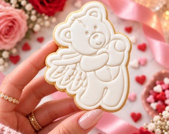 May include: A sugar cookie shaped like a teddy bear with angel wings, decorated with smooth white icing. The bear features embossed details including a stitched line on its head and chest, with a light brown baked edge visible around the perimeter.