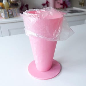 Pode incluir: Um suporte de saco de confeitar de plástico rosa com um saco de plástico transparente no interior. O suporte tem formato de cone e fica em uma base redonda. Fundo branco.