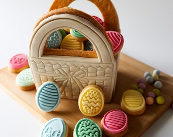 Op de afbeelding: Een witte keramische mand in de vorm van een handtas gevuld met kleurrijke paaseierenkoekjes. De mand is gemaakt van koekdeeg en staat op een houten oppervlak. Er liggen meer koekjes rond de mand, waaronder blauwe, groene, gele en roze eivormige koekjes.