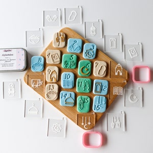 Op de afbeelding: Een set van 16 transparante koekjesuitstekers van plastic met verschillende islamitische ontwerpen, waaronder een halve maan, een moskee, een lantaarn en een boek. De uitstekers zijn gerangschikt op een houten plank met 16 kleurrijke koekjes die met dezelfde ontwerpen zijn versierd.