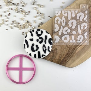 May include: A white fondant cookie decorated with a black leopard print design. A pink plastic cookie cutter with a cross design and a clear plastic stencil with a leopard print design are also shown.