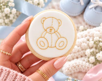 Könnte beinhalten: Ein runder Keks mit glatter weißer Glasur, der einen goldenen Umriss eines sitzenden Teddybären zeigt. Der Keks wird in der Hand gehalten, im Hintergrund sind hellblaue Babyschuhe und weiße Schleierkrautblüten zu sehen.