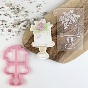 Könnte beinhalten: Ein rosa Plastik-Ausstecher in Form eines Kuchens mit floralen Details. Ein weißer Fondantkuchen mit rosa und grünen floralen Details befindet sich auf einer Holzoberfläche neben einer durchsichtigen Plastikschablone eines Kuchens mit floralen Details.