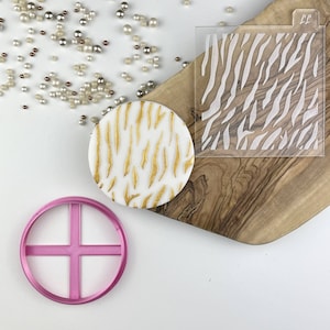 Pode incluir: Um bolo redondo de fondant branco com um design de zebra dourado. Também é mostrado um cortador de bolo de plástico rosa em forma de cruz. Uma plantilla de plástico transparente com um design de zebra branco é mostrada em uma superfície de madeira.