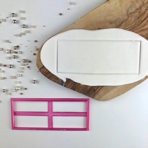 May include: A pink plastic cookie cutter with a rectangular shape divided into four sections by horizontal lines. The cutter is on a white surface.