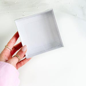 Puede incluir: Una pequeña caja cuadrada de cartón blanco con los lados plegados. La caja está sostenida por una mano con anillos en los dedos. La caja está vacía y parece nueva. El fondo es blanco.