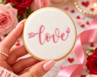 Puede incluir: Una galleta redonda con una base de glaseado blanco liso que presenta la palabra love escrita en cursiva rosa, con la letra l estilizada como una flecha que apunta a la derecha y la letra e terminando en punta de flecha.